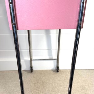May include: A vintage pink and chrome side table. The table has a rectangular pink painted wooden box supported by a chrome frame with wheels. The chrome frame has a rectangular top and bottom with curved legs.
