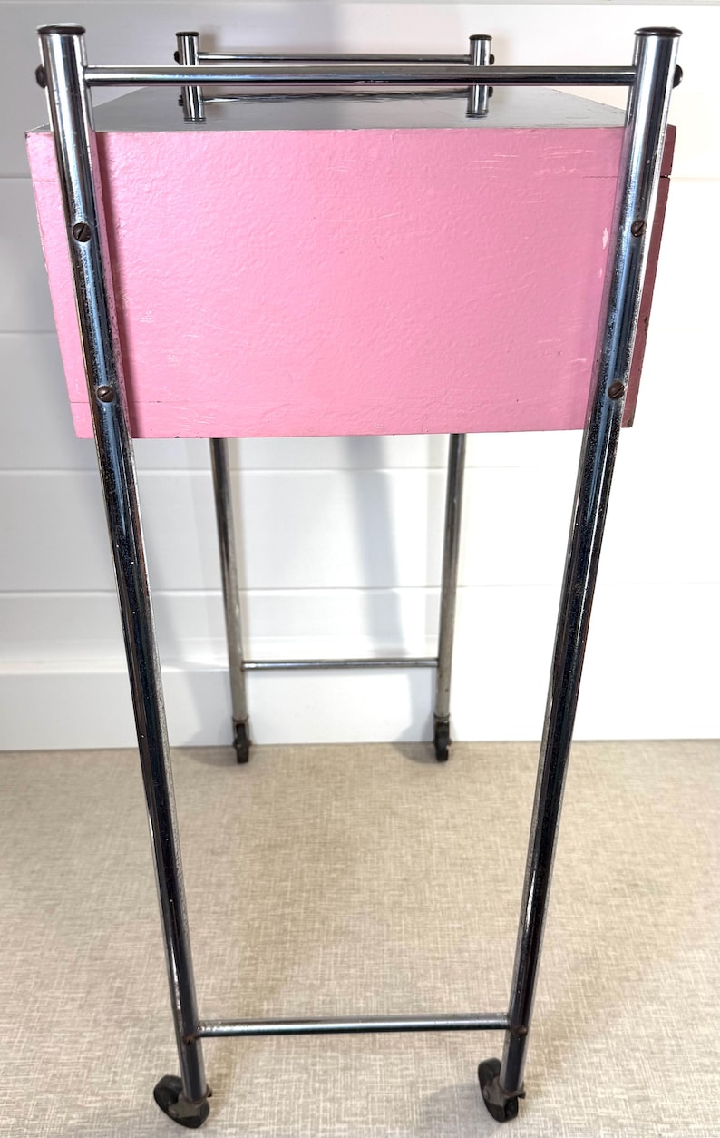 May include: A vintage pink and chrome side table. The table has a rectangular pink painted wooden box supported by a chrome frame with wheels. The chrome frame has a rectangular top and bottom with curved legs.