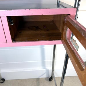 May include: A pink and gray rolling cart with a drawer and an open cabinet. The cart has a pink painted wooden frame, a gray top, and chrome legs with wheels. The cabinet door is open, revealing the wooden interior.