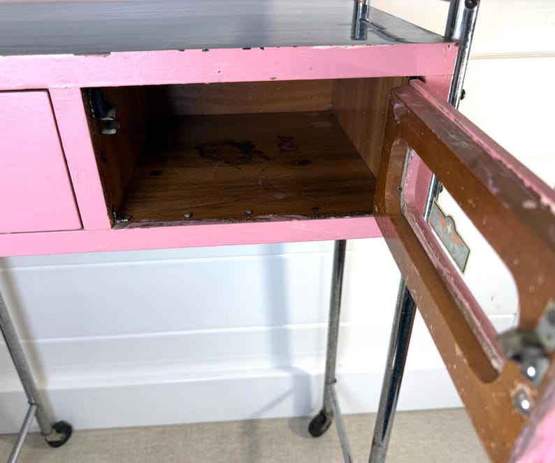 May include: A pink and gray rolling cart with a drawer and an open cabinet. The cart has a pink painted wooden frame, a gray top, and chrome legs with wheels. The cabinet door is open, revealing the wooden interior.
