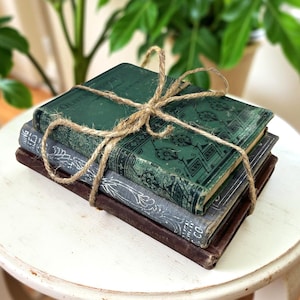 May include: A stack of three antique books tied with twine. The top book is green with ornate gold detailing, the middle book is gray, and the bottom book is brown. The books are stacked on a white, round table.