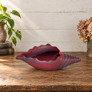 May include: A conch shell-shaped bowl with a matte finish in deep burgundy and muted blue tones. The bowl features a wide, undulating opening and a tapered, spiraled tail, showcasing a smooth, solid-colored surface throughout its entire form.