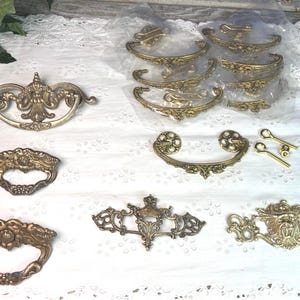 May include: A collection of antique brass drawer pulls and handles. The hardware features ornate, detailed designs with a vintage aesthetic. Various shapes and sizes are displayed, including pulls with keyhole details and decorative backplates.