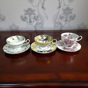 May include: Three decorative teacups and saucers are arranged on a dark wooden surface. The teacups feature floral designs in various colors, including pink, yellow, and green. Each cup sits on a matching saucer, creating a classic tea set.