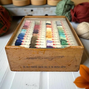 May include: A vintage wooden thread box with a glass lid, filled with colorful spools of thread. The box is labeled "The Most Modern Thread Mill in the British Empire." Several balls of yarn in various colors are in the background.