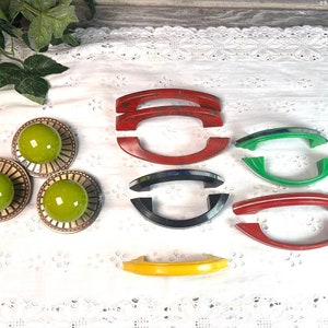 May include: Assortment of vintage cabinet knobs and drawer pulls in various colors, including red, green, yellow, and black. The knobs are round with a green glass center and a brass rim. The pulls are made of plastic and have a curved shape.