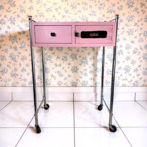 May include: A vintage pink bedside table with two drawers and a cabinet door. The table has a chrome frame and sits on four small wheels. The drawers have black handles, and the cabinet door has a black lock. The background is a floral wallpaper.
