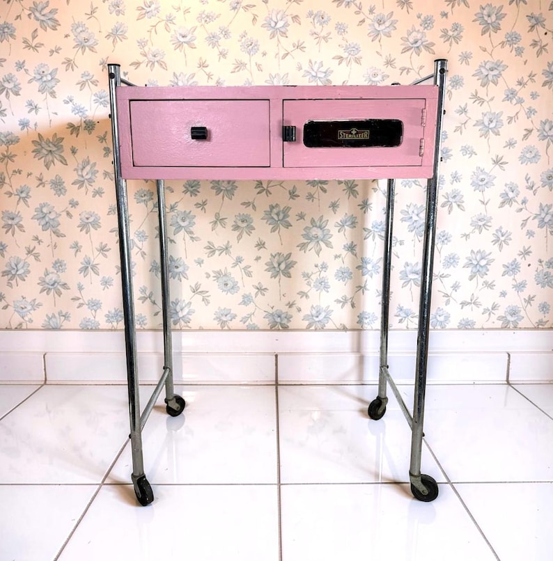 May include: A vintage pink bedside table with two drawers and a cabinet door. The table has a chrome frame and sits on four small wheels. The drawers have black handles, and the cabinet door has a black lock. The background is a floral wallpaper.