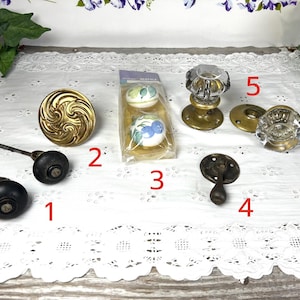 May include: Assortment of ten antique door knobs and pulls in various styles and finishes, including brass, glass, and black metal. The knobs feature a variety of designs, including a swirl pattern, a clear glass knob, and a black metal knob with a simple design.