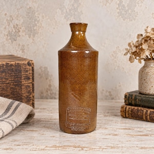 May include: A tall, cylindrical brown stoneware bottle with a narrow neck and a flared rim. The front features an embossed rectangular stamp with the text: LIQUEFIED STONE BOTTLE, J. BOURNE & SON, DENBY POTTERY, NEAR DERBY, P. & J. ARNOLD, LONDON, ENGLAND.