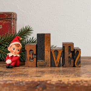 May include: A small elf figurine wearing a red hat and red clothing sits beside a set of vintage wooden letterpress blocks arranged to spell the word ELVES. The blocks are brown with black ink residue and have varying heights and widths.