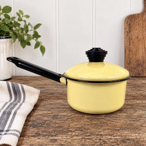 May include: A small, pale yellow enamel saucepan with a black handle and a matching lid featuring a tiered black knob. The pot has a smooth, glossy finish and a black rim, designed for stovetop use with a single long handle for easy gripping.
