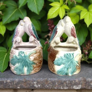 May include: Two ceramic bird planters. Each planter features a pair of kissing birds perched above a small opening. The planters are painted in shades of white, green, brown, and purple. A large green leaf is on the front of each planter.