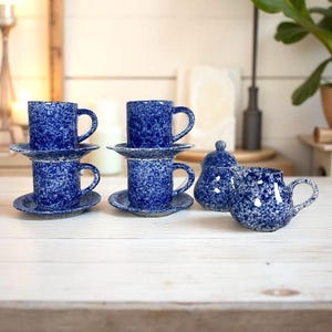 May include: A set of blue speckled ceramic teacups and saucers, a creamer, and a sugar bowl with a lid. The teacups are stacked in pairs. The set is on a white wooden surface.