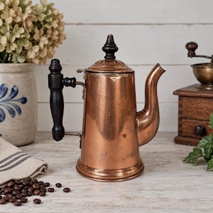 May include: A copper coffee pot featuring a cylindrical body, a curved spout, and a black wooden handle attached to the side. The lid is topped with a black wooden finial. The metal surface shows a warm, metallic finish with subtle signs of wear.