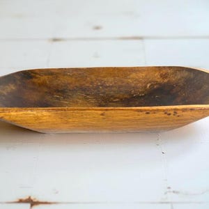 May include: A rustic, wooden dough bowl with a natural, aged appearance. The bowl is oval-shaped with a shallow depth and a warm, golden-brown color. The interior shows darker tones, adding to its vintage charm.
