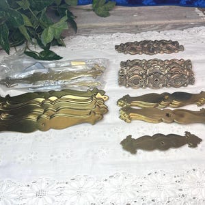 May include: An assortment of vintage brass hardware, including drawer pulls and decorative accents. The pieces vary in size and design, with ornate scrollwork and a warm, golden hue. Some are still in their original packaging.