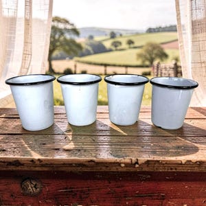 May include: Four white enamel cups with black rims are arranged on a weathered wooden surface. The cups are empty and have a slightly distressed appearance. The background features a scenic view of rolling hills and a bright sky.