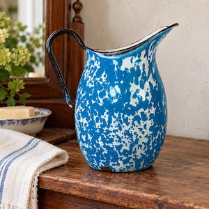 May include: A blue and white speckled enamel pitcher with a curved black handle and a wide pouring spout. The exterior features a mottled pattern of blue paint over a white base, with a dark rim and a small area of wear near the base.