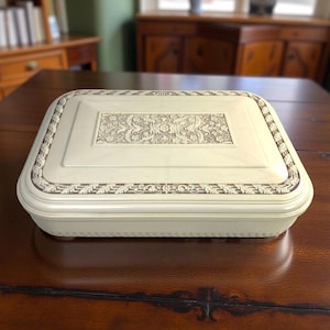 May include: A rectangular, off-white jewelry box with a decorative floral design on the lid. The box has a raised, ornate central panel and a detailed border. The box is sitting on a dark brown wooden table.