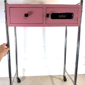 May include: A vintage pink cabinet with a drawer and a door, mounted on a chrome rolling frame. The cabinet has a black handle and the word "STERILIZER" is visible on the door.