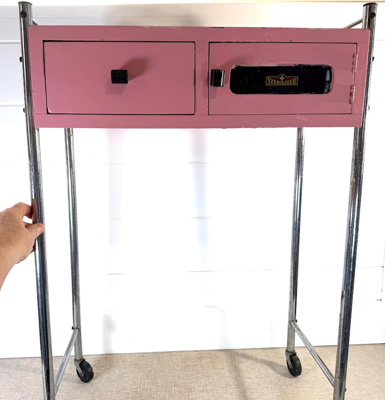 May include: A vintage pink cabinet with a drawer and a door, mounted on a chrome rolling frame. The cabinet has a black handle and the word "STERILIZER" is visible on the door.