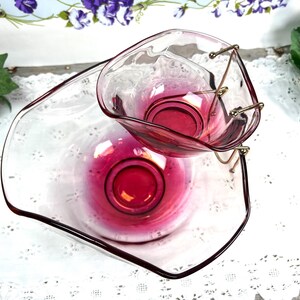 May include: A vintage cranberry-colored glass serving set. The set includes a large, shallow dish and a smaller, heart-shaped dish held by a gold-toned metal stand. The glass has a glossy finish, and the set is likely used for serving snacks or appetizers.