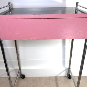 May include: A vintage pink and chrome rolling cart. The cart has a rectangular pink painted drawer and a black top. The frame is made of chrome and has small wheels on the bottom for easy movement. The cart is likely from the mid-century era.
