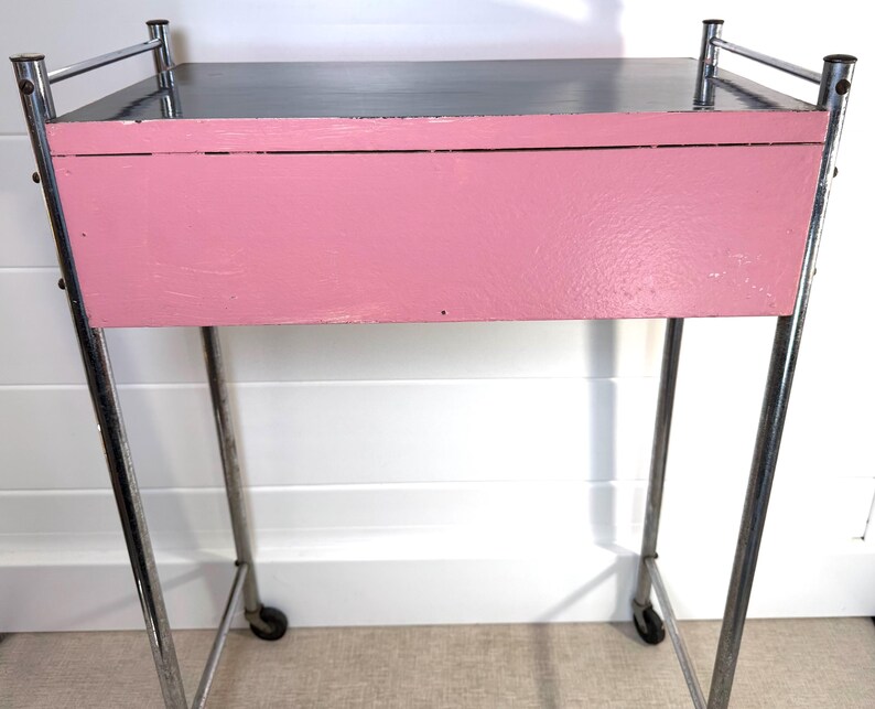 May include: A vintage pink and chrome rolling cart. The cart has a rectangular pink painted drawer and a black top. The frame is made of chrome and has small wheels on the bottom for easy movement. The cart is likely from the mid-century era.