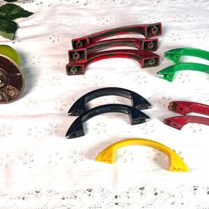May include: Assortment of vintage cabinet knobs in various colors, including red, green, black, and yellow. The knobs are made of plastic and have a rounded, arched shape.