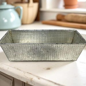 May include: A rectangular, galvanized metal baking pan with a textured surface. The pan has angled sides and is empty, sitting on a white surface. A light blue teapot is visible in the background, suggesting a kitchen setting.