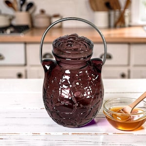 May include: A dark purple glass jar with a lid and a metal handle. The jar has a textured floral design. A small glass bowl with honey and a wooden spoon are next to the jar. The background is a kitchen.