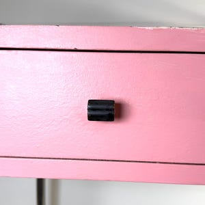May include: Close-up of a pink painted drawer with a black handle. The drawer is part of a larger piece of furniture, possibly a desk or a shelf. The paint has a slightly distressed look.