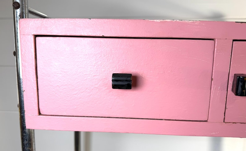 May include: Close-up of a pink painted drawer with a black handle. The drawer is part of a larger piece of furniture, possibly a desk or a shelf. The paint has a slightly distressed look.