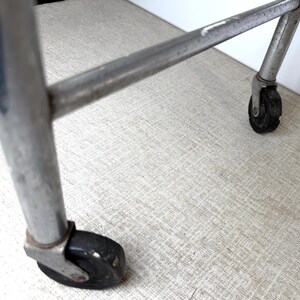 May include: Close-up of a chrome metal cart frame with black wheels. The wheels are small and round, and the frame is slightly angled. The background is a light gray textured surface.