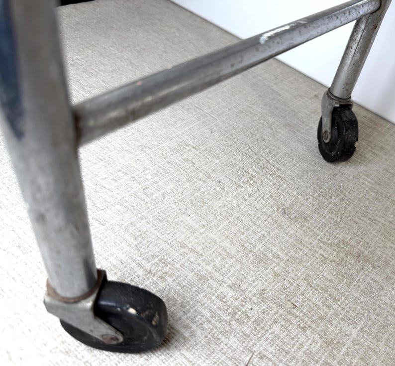 May include: Close-up of a chrome metal cart frame with black wheels. The wheels are small and round, and the frame is slightly angled. The background is a light gray textured surface.