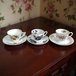 May include: Three antique teacups and saucers with floral designs sit on a dark wood surface. The cups and saucers feature delicate patterns in shades of purple, yellow, and pink, with gold trim. The background is a floral wallpaper.