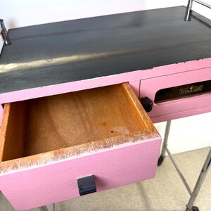May include: A pink and black painted wooden cart with a drawer open. The drawer is made of wood and has a black handle. The cart has chrome bars and wheels.