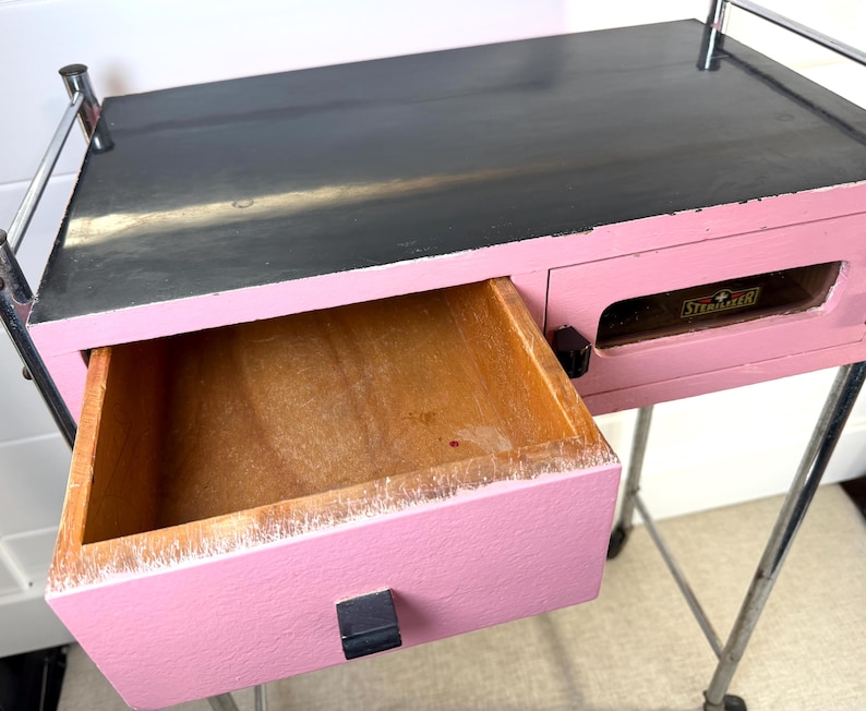 May include: A pink and black painted wooden cart with a drawer open. The drawer is made of wood and has a black handle. The cart has chrome bars and wheels.