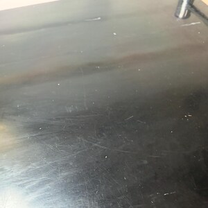 May include: A close-up shot of a black, glossy surface with visible scratches and imperfections. The surface appears to be part of a larger object, possibly a table or countertop. The lighting creates highlights and shadows.