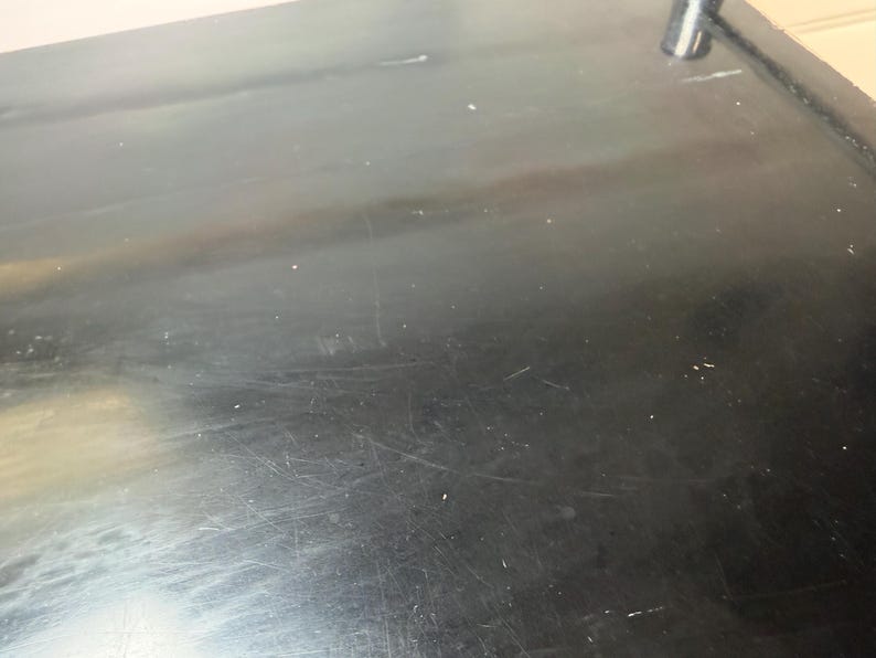 May include: A close-up shot of a black, glossy surface with visible scratches and imperfections. The surface appears to be part of a larger object, possibly a table or countertop. The lighting creates highlights and shadows.