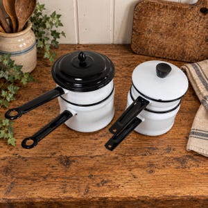 May include: Two double boiler sets made of white enamel with black rims and black handles. One set features a solid black lid, while the other has a white lid with a black knob. Each pot has a long handle with a circular hole at the end.