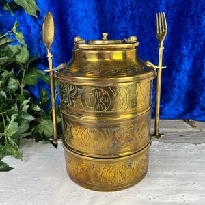 May include: A three-tiered brass tiffin carrier with intricate engraved designs. The carrier has two attached utensils, a spoon and a fork.