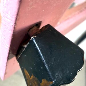 May include: Close-up of a black, square-shaped furniture leg with visible wear and tear, attached to a pink wooden structure. The leg shows signs of age with exposed wood and rust-colored discoloration.