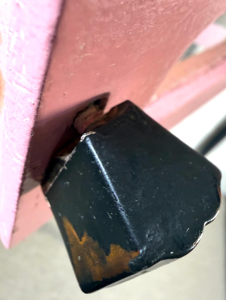 May include: Close-up of a black, square-shaped furniture leg with visible wear and tear, attached to a pink wooden structure. The leg shows signs of age with exposed wood and rust-colored discoloration.