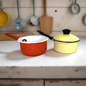 May include: Two vintage enamelware pots. One is bright orange with a white interior and a red handle. The other is yellow with a black handle and lid. Kitchen utensils hang in the background.