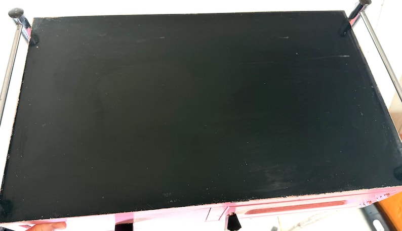 May include: A black, rectangular tabletop with a glossy finish. The table is supported by a metal frame with chrome-colored bars. The edges of the table are pink.
