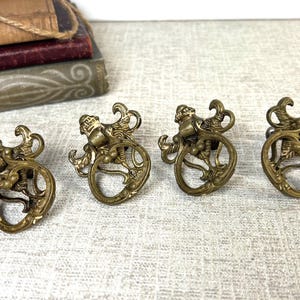 May include: Four antique brass drawer pulls with a detailed design featuring a figure atop a decorative ring. The pulls are a warm, aged brass color, suggesting vintage charm. A stack of old books is in the background.