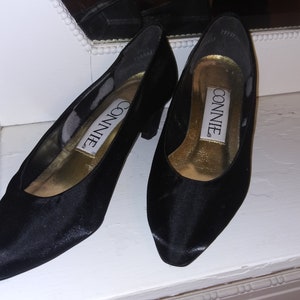 May include: A pair of black satin pumps with a pointed toe and a low heel. The shoes are labeled "Connie" on the inside.
