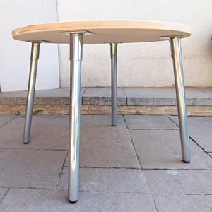 Könnte beinhalten: Ein runder Holztisch mit vier silbernen Metallbeinen. Der Tisch steht auf einem Fliesenboden.